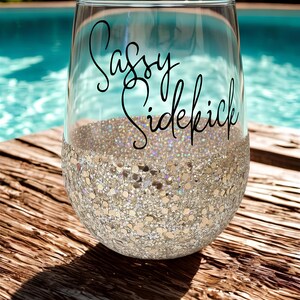 A clear wine glass with a glitter-covered base. The glass has the words "Sassy Sidekick" in black script. The glass can be personalized with a name or slogan