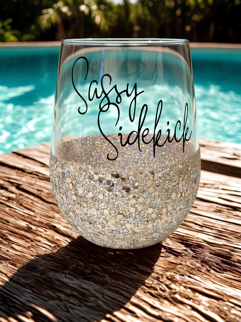 A clear wine glass with a glitter-covered base. The glass has the words "Sassy Sidekick" in black script. The glass can be personalized with a name or slogan