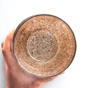 A clear glass with a textured, speckled interior. The glass has a metallic gold and brown color scheme, with a rim that appears to be a darker shade of gold. The glass is held by a person, and the background is white.