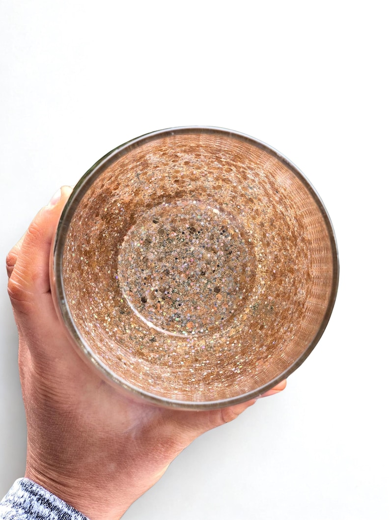 A clear glass with a textured, speckled interior. The glass has a metallic gold and brown color scheme, with a rim that appears to be a darker shade of gold. The glass is held by a person, and the background is white.