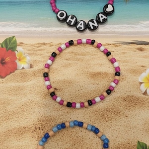 Puede incluir: Tres pulseras circulares de cuentas. La pulsera superior tiene cuentas rosas y cuentas con letras negras que forman la palabra OHANA con un coraz&oacute;n blanco. La pulsera central tiene un patr&oacute;n de cuentas rosas, blancas, negras y color canela. La pulsera inferior tiene cuentas azules y color canela.