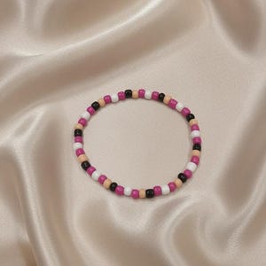 Puede incluir: Una pulsera circular de cuentas que presenta un patr&oacute;n repetitivo de peque&ntilde;as cuentas esf&eacute;ricas y lisas en colores alternos negro, blanco, rosa magenta y un tono beige suave, dispuestas en un bucle continuo sobre una superficie lisa.