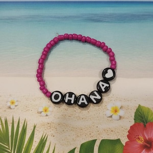 Puede incluir: Una pulsera circular hecha de peque&ntilde;as cuentas de color magenta brillante. La pulsera presenta cuentas negras, redondas y planas con letras blancas que forman la palabra OHANA, seguidas de una cuenta con forma de coraz&oacute;n blanco al final.