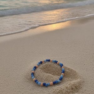 Puede incluir: Una pulsera circular compuesta por peque&ntilde;as cuentas lisas en tonos alternos de azul oscuro, azul claro y melocot&oacute;n suave. Las cuentas est&aacute;n dispuestas en un patr&oacute;n repetitivo, creando un dise&ntilde;o de bucle simple y continuo sobre la arena.