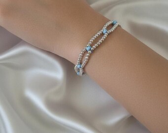 Pulsera de doble hebra con cuentas plateadas y cuentas azul claro, unida mediante un cierre de mosquetón plateado.