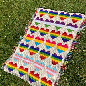 May include: A crocheted blanket with a white background and rainbow hearts. The blanket has a fringe border.