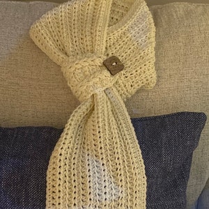 May include: Cream-colored, hand-crocheted scarf with a fringed edge. The scarf features a textured stitch pattern and is secured with a square, wooden button. The scarf is draped over a blue pillow on a gray couch.