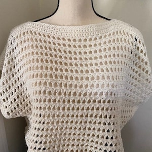 May include: Off-white, open-weave crochet top with a boat neck and short sleeves. The top is made of a thick yarn, creating a textured, airy design. The garment is displayed on a mannequin.