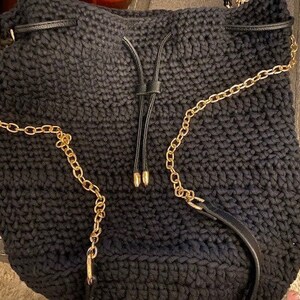 May include: Black crochet shoulder bag with gold chain straps and a drawstring closure. The bag has a textured appearance and a leather strap detail. The gold chain adds a touch of elegance to the casual design.