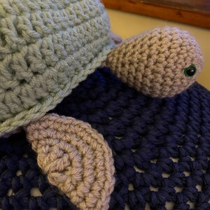 May include: A handmade crochet turtle hat in light blue with tan-colored flippers and head. The turtle has a green eye. The hat is resting on a dark blue crocheted surface. The hat is a unique, handmade item.