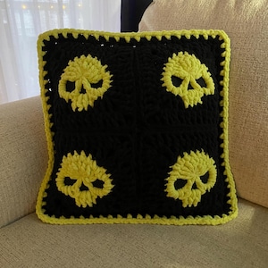 May include: A square, handmade crochet pillow featuring four yellow skull designs on a black background. The pillow has a yellow border and is placed on a beige sofa. The skulls are a prominent design element.