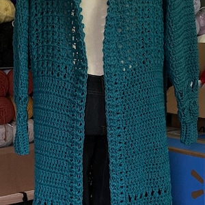 May include: A long, teal-colored, open-front crochet cardigan with a hood. The handmade garment features a textured stitch pattern and decorative cutouts near the hem and sleeves. The cardigan is displayed on a mannequin.