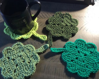 May include: Four crocheted shamrock-shaped coasters in shades of green. One coaster is under a black coffee mug. The coasters are handmade and have a textured appearance, perfect for St. Patrick's Day or everyday use.