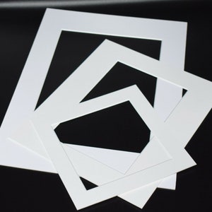 May include: A collection of white picture mats in various sizes, arranged on a black surface. The mats have rectangular openings, designed for framing artwork or photographs. The clean, white mats provide a crisp, professional look.