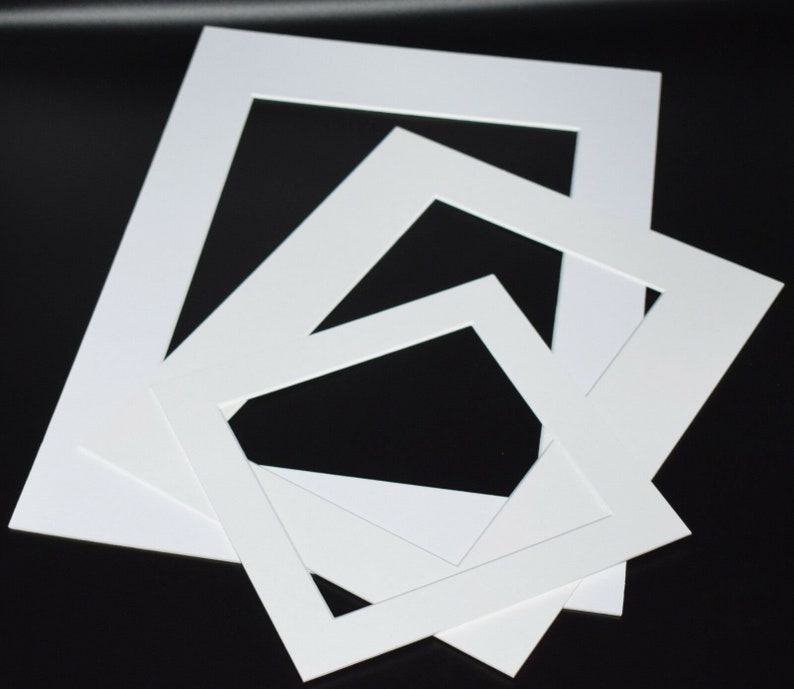 May include: A collection of white picture mats in various sizes, arranged on a black surface. The mats have rectangular openings, designed for framing artwork or photographs. The clean, white mats provide a crisp, professional look.