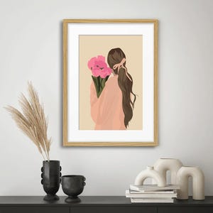 May include: A framed wall art print featuring a digital illustration of a person with long brown hair tied with a pink ribbon, wearing a peach-colored top and holding a bouquet of bright pink flowers against a solid light beige background.
