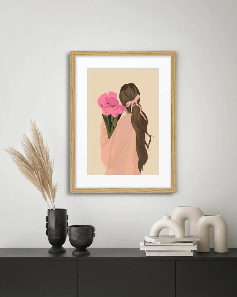 May include: A framed wall art print featuring a digital illustration of a person with long brown hair tied with a pink ribbon, wearing a peach-colored top and holding a bouquet of bright pink flowers against a solid light beige background.