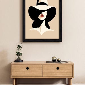 Peut inclure: Une impression d'art encadr&eacute;e repr&eacute;sentant une illustration stylis&eacute;e d'une personne portant un chapeau cr&egrave;me &agrave; larges bords avec un ruban noir. La silhouette a des cheveux noirs, un teint cr&egrave;me et des l&egrave;vres rouge vif sur fond beige clair.