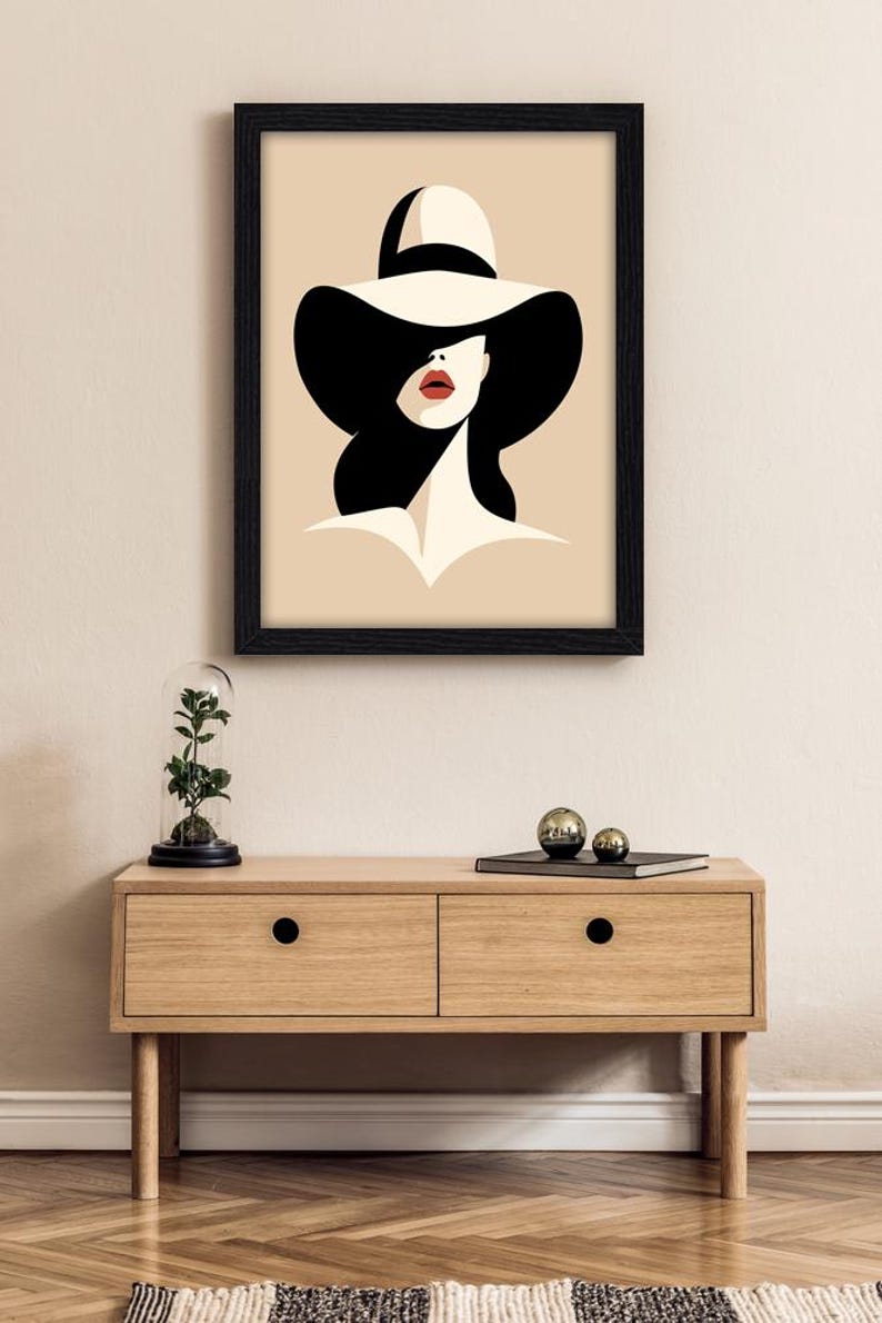 Peut inclure: Une impression d'art encadr&eacute;e repr&eacute;sentant une illustration stylis&eacute;e d'une personne portant un chapeau cr&egrave;me &agrave; larges bords avec un ruban noir. La silhouette a des cheveux noirs, un teint cr&egrave;me et des l&egrave;vres rouge vif sur fond beige clair.