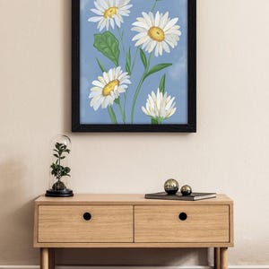 Peut inclure: Une impression d'art encadr&eacute;e repr&eacute;sentant une illustration botanique de marguerites blanches aux centres jaunes et aux feuilles vertes sur un fond bleu nuageux. L'&oelig;uvre est pr&eacute;sent&eacute;e dans un cadre rectangulaire noir simple.