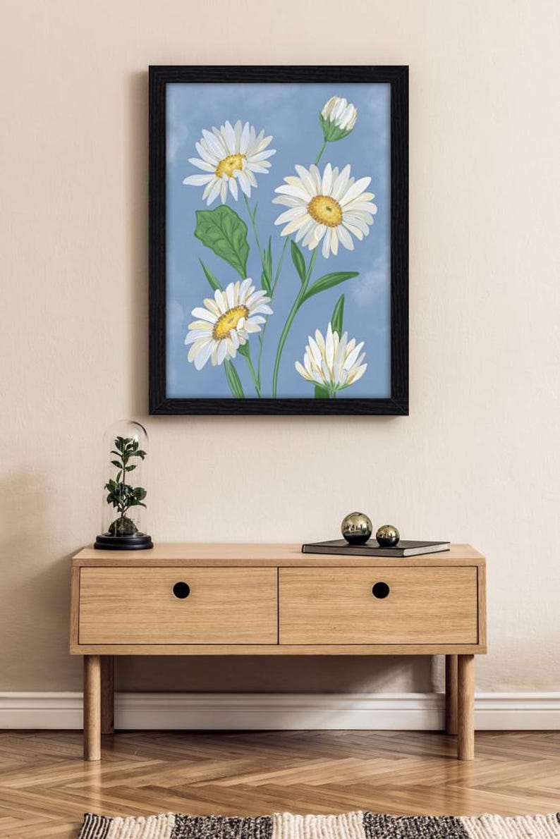 Peut inclure: Une impression d'art encadr&eacute;e repr&eacute;sentant une illustration botanique de marguerites blanches aux centres jaunes et aux feuilles vertes sur un fond bleu nuageux. L'&oelig;uvre est pr&eacute;sent&eacute;e dans un cadre rectangulaire noir simple.