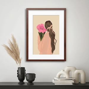 May include: A framed wall art print featuring a digital illustration of a person with long brown hair tied with a pink ribbon, wearing a peach-colored top and holding a bouquet of bright pink flowers against a solid light beige background.