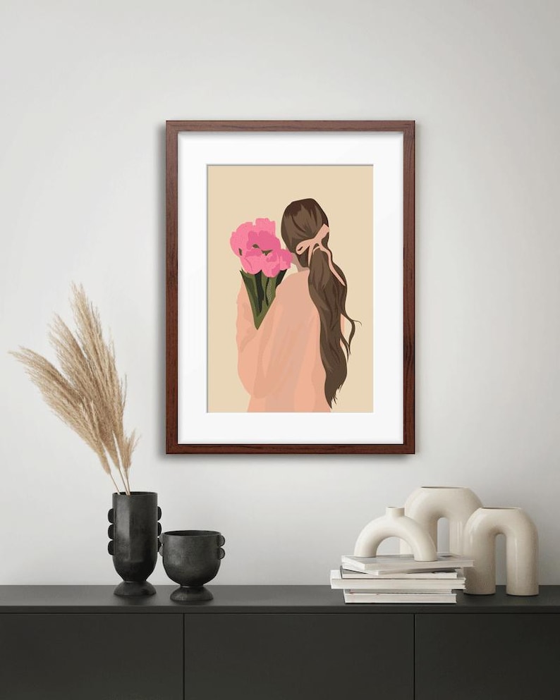 May include: A framed wall art print featuring a digital illustration of a person with long brown hair tied with a pink ribbon, wearing a peach-colored top and holding a bouquet of bright pink flowers against a solid light beige background.