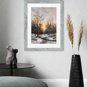 May include: A framed wall art print featuring a winter landscape painting with bare trees, snow-covered ground, a small stream, and a lone deer under a soft orange and blue sunset sky, enclosed in a weathered light gray wooden frame with a white mat.