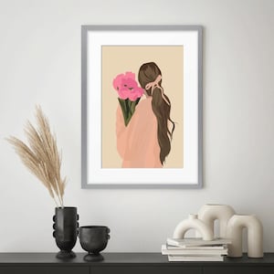 May include: A framed wall art print featuring a digital illustration of a person with long brown hair tied back with a ribbon, wearing a peach-colored top and holding a bouquet of bright pink flowers against a solid light beige background.