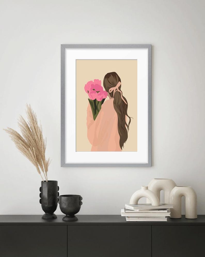 May include: A framed wall art print featuring a digital illustration of a person with long brown hair tied back with a ribbon, wearing a peach-colored top and holding a bouquet of bright pink flowers against a solid light beige background.