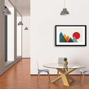 May include: A framed wall art print featuring a geometric mountain landscape with overlapping triangles in shades of blue, green, orange, and yellow, set against a cream background with a large solid red circle representing a sun in the sky.