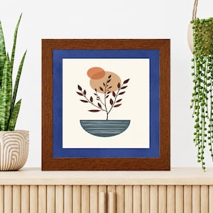 May include: A square wall art print in a dark brown wooden frame with a blue mat. The print features a stylized dark brown plant with leaves growing from a blue semi-circular bowl, set against a cream background with two overlapping tan circles.