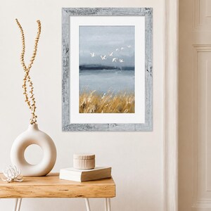 May include: A framed wall art print featuring a landscape painting of a flock of white birds flying over a calm blue body of water with golden reeds in the foreground. The artwork is enclosed in a distressed white wooden frame with a white mat.
