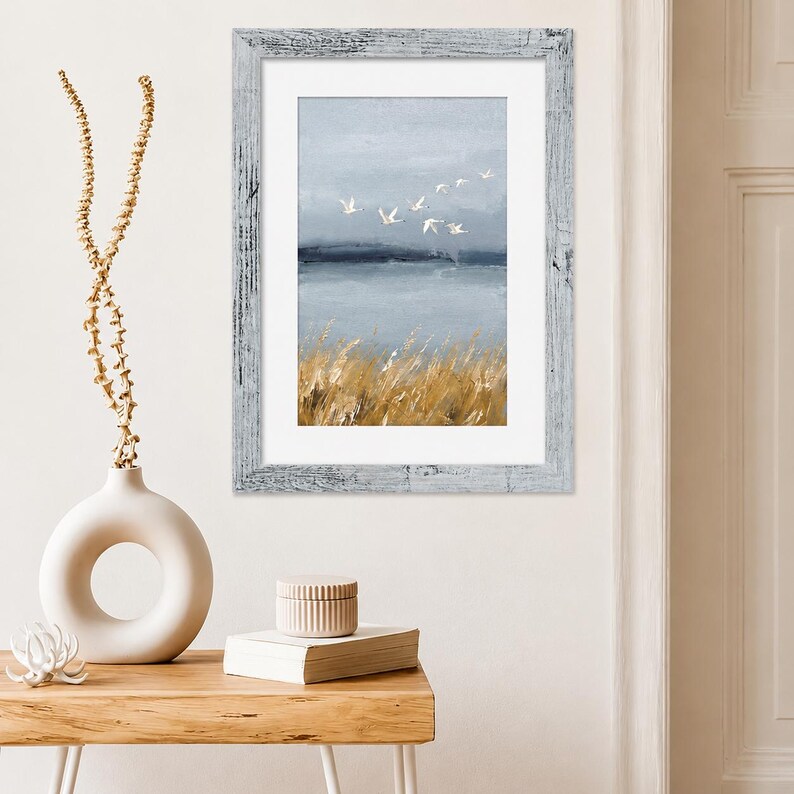May include: A framed wall art print featuring a landscape painting of a flock of white birds flying over a calm blue body of water with golden reeds in the foreground. The artwork is enclosed in a distressed white wooden frame with a white mat.