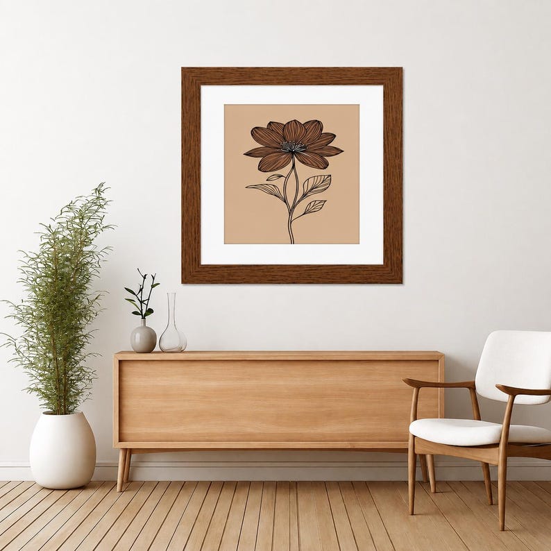 May include: A square framed wall art piece featuring a black line drawing of a single flower with detailed petals and leaves centered on a solid tan background. The artwork is enclosed in a dark brown wooden frame with a white mat border.