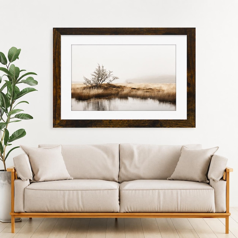 May include: A framed landscape photograph featuring a lone tree on a grassy hill above a reflective body of water, set in a dark wood frame with a white mat. The scene is displayed above a beige sofa with two matching throw pillows.