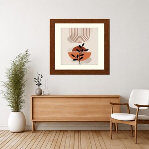 May include: A square framed wall art piece featuring a beige background with a series of thin, dark red concentric arches at the top, a solid terracotta semi-circle in the center, and a black silhouette of a leafy plant stem layered over it.