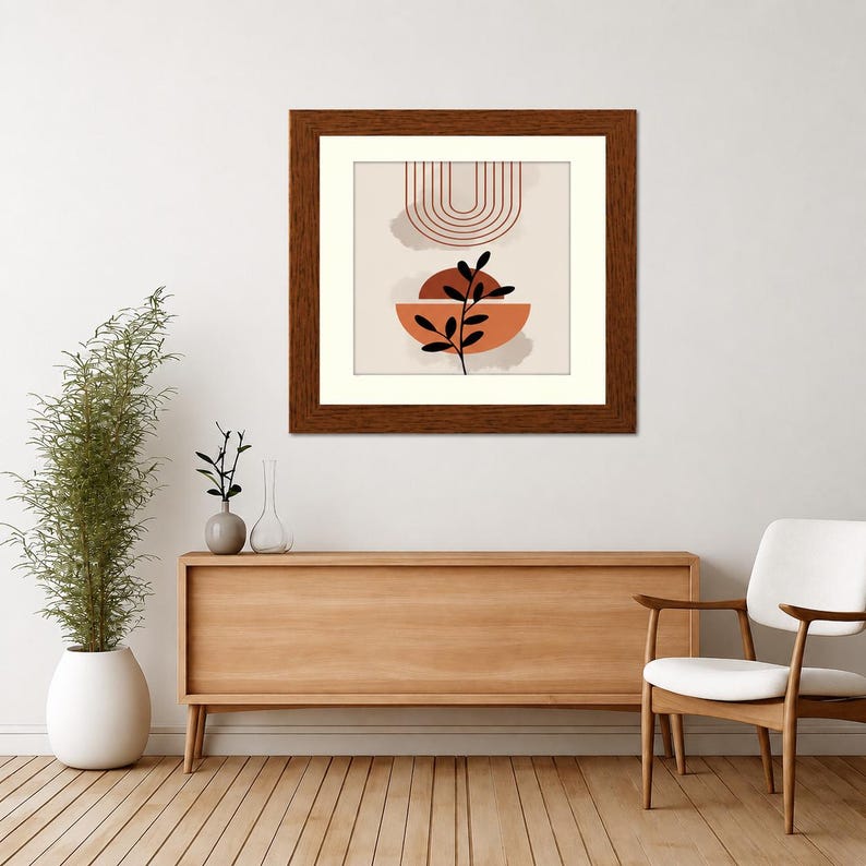 May include: A square framed wall art piece featuring a beige background with a series of thin, dark red concentric arches at the top, a solid terracotta semi-circle in the center, and a black silhouette of a leafy plant stem layered over it.