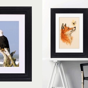 May include: Two black rectangular frames with white matting. The left frame displays a bald eagle perched on a weathered tree stump against a blue sky and ocean. The right frame contains a watercolor illustration of a fox looking at a small bee.