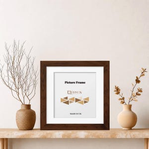 May include: A square dark brown wooden picture frame with a white mount. The insert features the text Picture Frame, the logo OSUK, a geometric pattern of triangles, and the text MADE IN UK. The frame is positioned between two decorative vases.