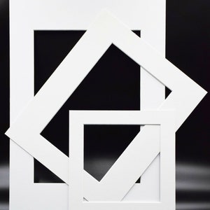 May include: Three white picture frame mats of varying sizes are arranged against a black background. The mats have square and rectangular openings, with one angled across the frame. The mats are clean and modern.