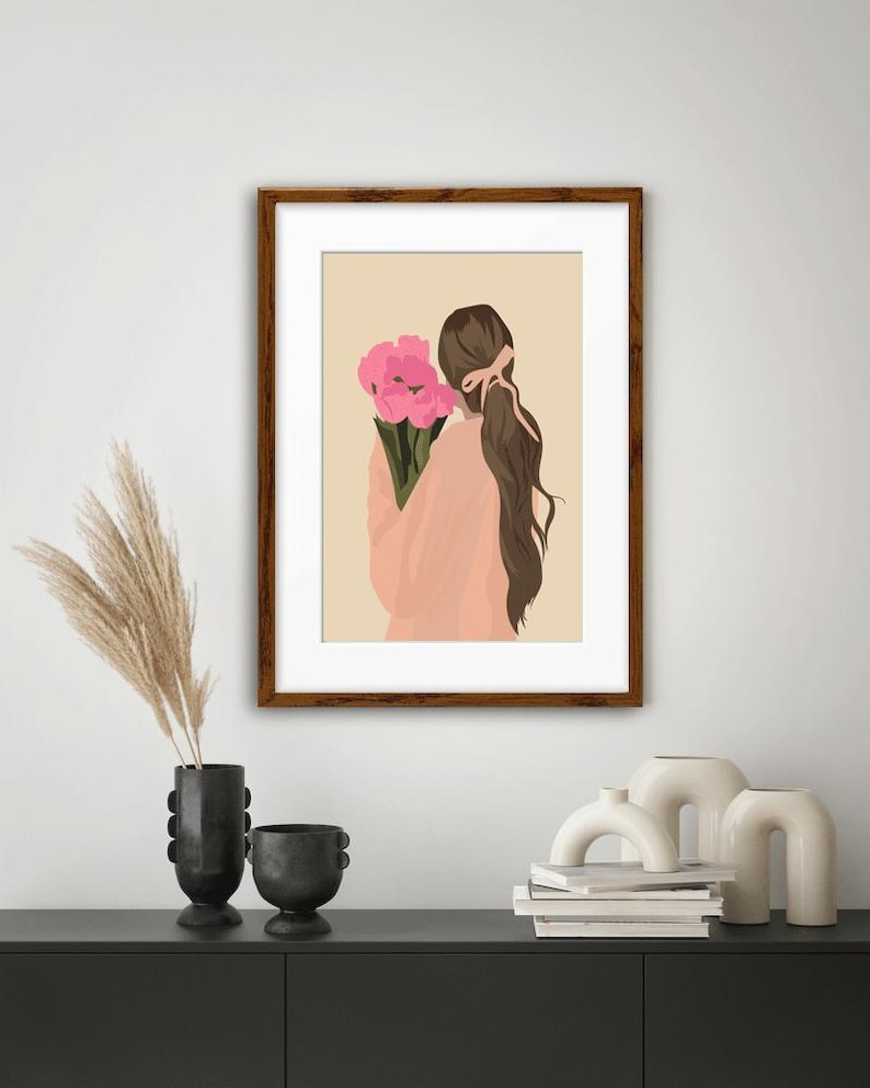 May include: A framed wall art print featuring a digital illustration of a person with long brown hair tied back with a ribbon, wearing a peach-colored top and holding a bouquet of bright pink flowers against a solid light beige background.