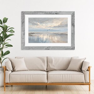 May include: A framed landscape painting depicting a serene coastal scene with a soft, cloudy sky, calm water, and distant birds in flight. The artwork is encased in a weathered, light gray wooden frame with a wide white mat border.