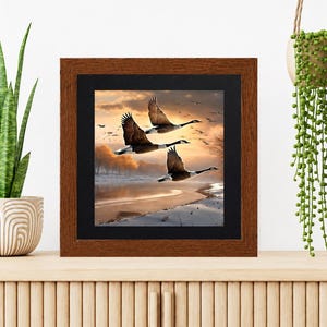 May include: A square wall art piece featuring a print of three geese in flight over a snowy landscape at sunset, encased in a dark brown wooden frame with a black mat border. The artwork depicts a golden sky and a frozen riverbank scene.