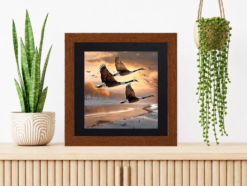 May include: A square wall art piece featuring a print of three geese in flight over a snowy landscape at sunset, encased in a dark brown wooden frame with a black mat border. The artwork depicts a golden sky and a frozen riverbank scene.
