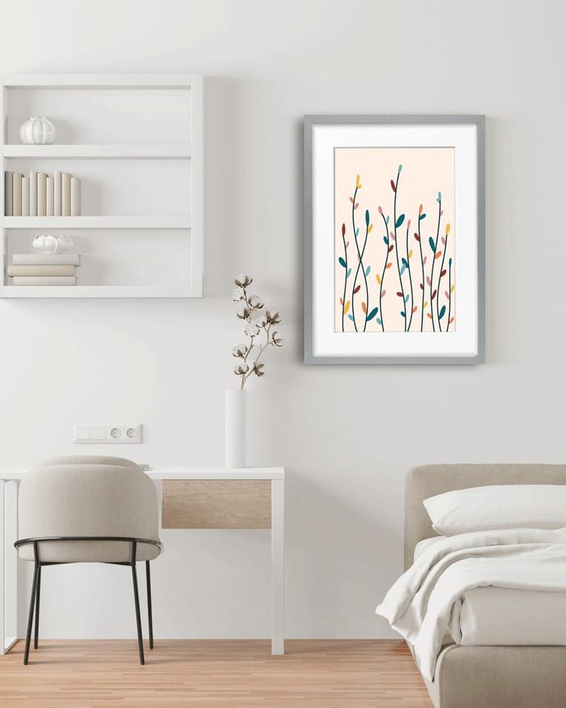 May include: A framed wall art print featuring a light peach background with vertical, slender stems and small, multi-colored leaves in shades of teal, yellow, pink, and dark brown. The artwork is enclosed in a simple, light gray rectangular frame.