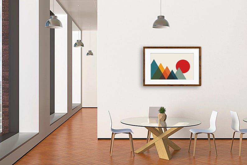 May include: A framed wall art print featuring a geometric mountain landscape with overlapping triangles in shades of blue, green, and orange, set against a cream background with a large, solid red circle representing a sun in the upper right corner.