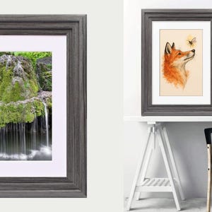 May include: A dark gray wooden picture frame with a white mat board. The frame features a stepped profile and holds a vertical rectangular print of a moss-covered rock formation with water cascading down into a pool at the base of the structure.