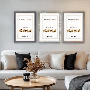May include: Three rectangular picture frames with the text "Picture Frame" and "MADE IN UK". The frames are gray, white, and light brown. The frames are hanging above a light grey sofa with black and white cushions.