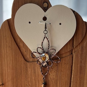 May include: A decorative pendant featuring a central clear faceted crystal surrounded by a flower-shaped frame made of small brown and metallic beads. A single faceted amber-colored bead hangs from the bottom of the beaded flower structure.